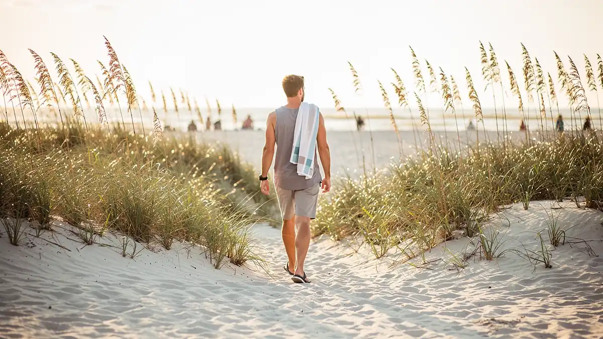 Crossing pale sand past sea oats with a towel over one shoulder