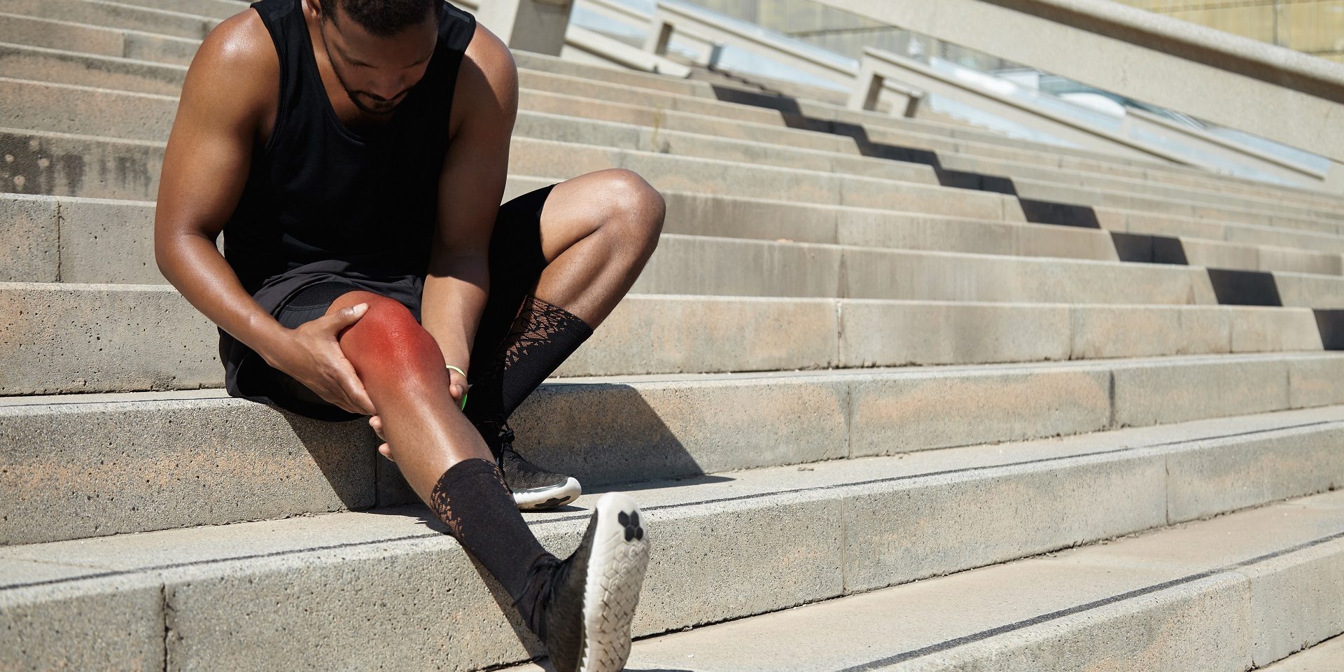 Attractive dark-skinned male runner wearing black outfit sitting on stairs outdoors, massaging his aching knee of red color, looking at sore skin with painful expression, trying to soothe pain Treatment Options for Sports-Related Foot Injuries